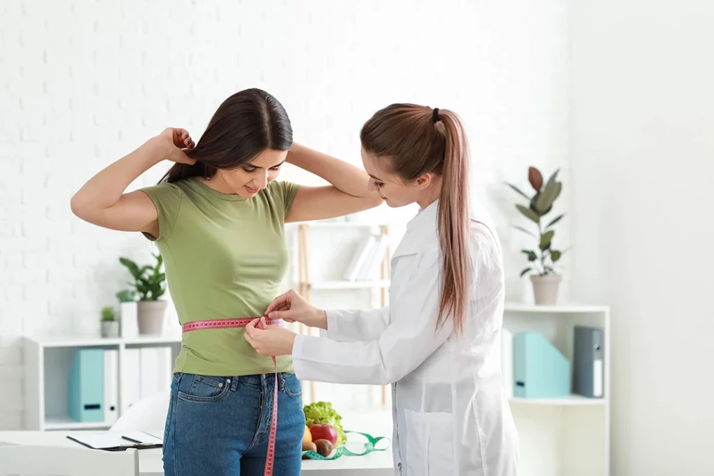 Nutritionist measures woman's waist with tape near fresh produce in consultation at weight loss clinic Houston.