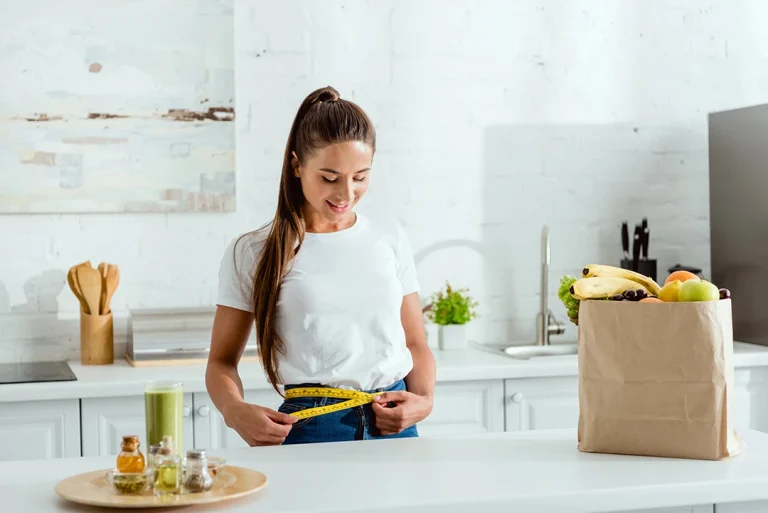 A woman measuring her waist with a tape in a kitchen, promoting semaglutide in Houston, with healthy groceries nearby.