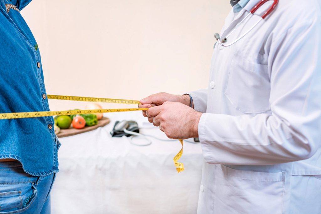 Expert weight loss doctor in Houston measuring patient's waist with tape during consultation, featuring fresh fruits, vegetables, and medical tools on table for healthy lifestyle guidance.