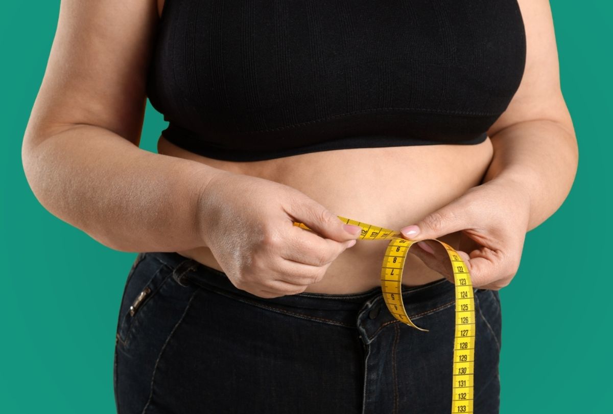 Individual in dark clothing measuring waist with yellow tape against teal background, illustrating weight loss monitoring