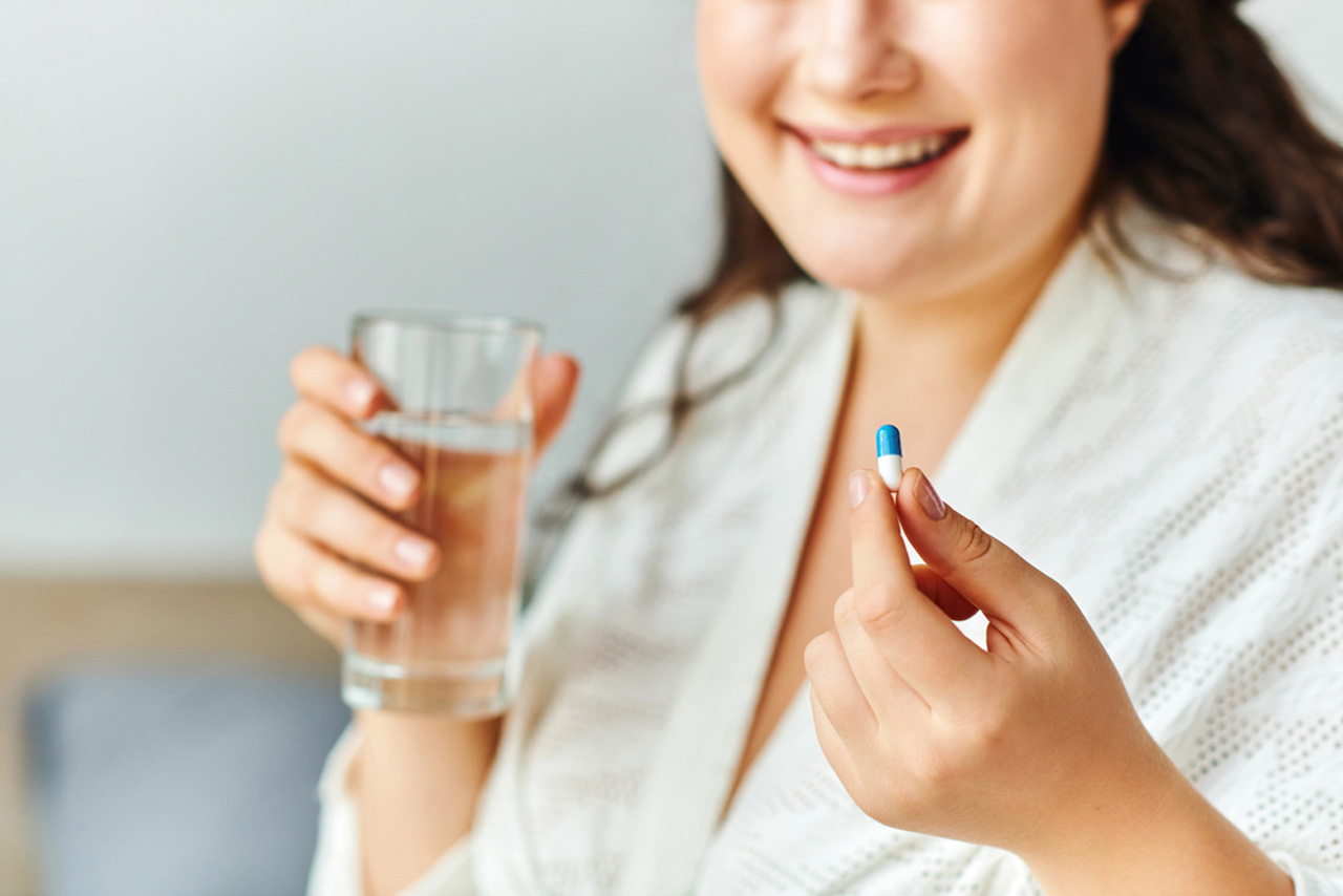 Smiling woman taking a weight loss pill with water at home, representing supervised Medical Weight Loss Houston support and guidance.