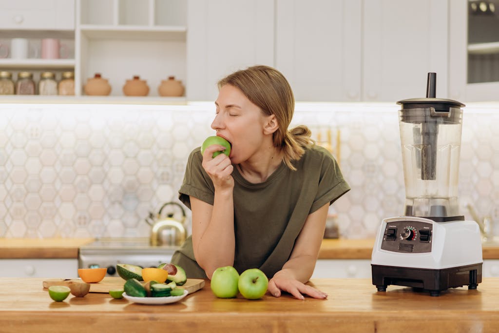 A woman bites into a fresh green apple in a modern kitchen setting, promoting healthy eating.
