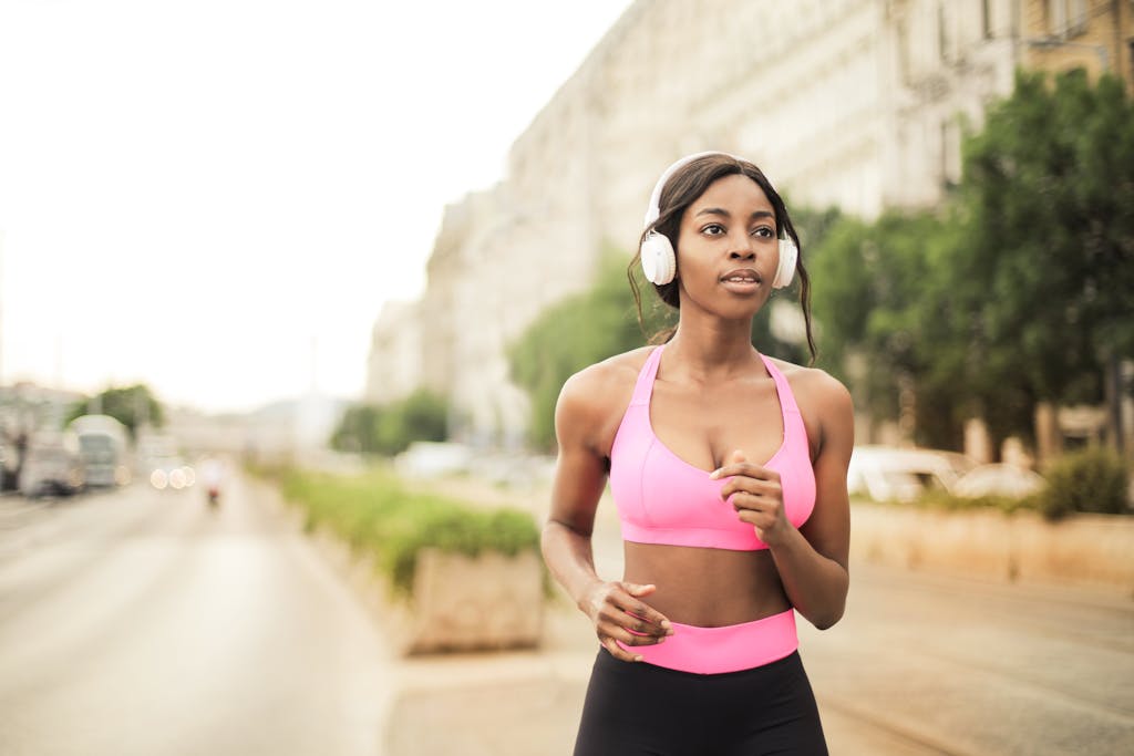Black woman jogging in city street wearing pink activewear and headphones.