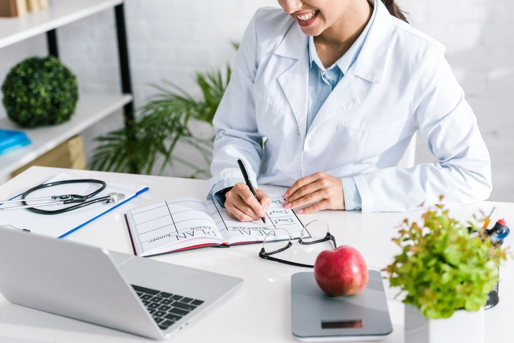 A smiling weight loss doctor Houston creating a personalized meal plan in a notebook at her clinic desk with a laptop.