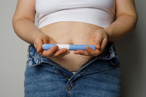 A person in a white top and unbuttoned jeans holding the injection pen horizontally across their midsection.