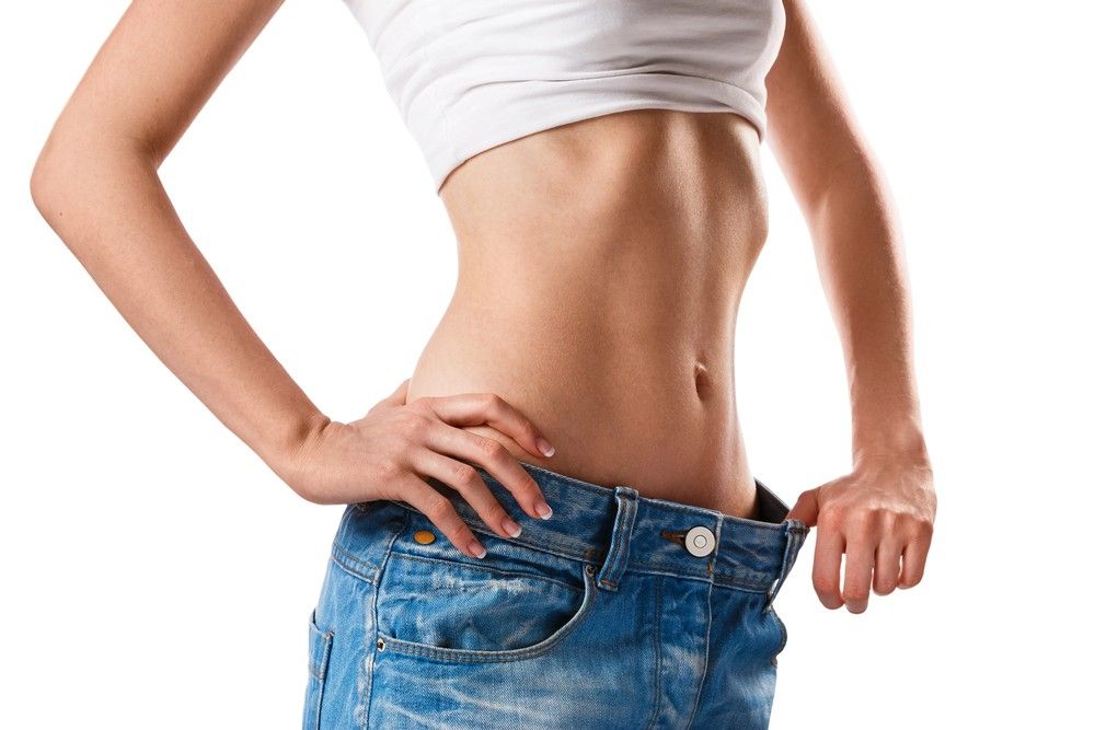 Fit woman in white crop top and light blue loose jeans has hands on hips after weight loss clinic Houston help.