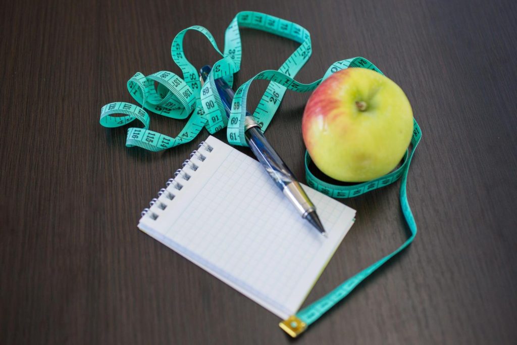 Tracking progress with Mounjaro for weight loss Houston using a notebook, apple, and measuring tape on a table.