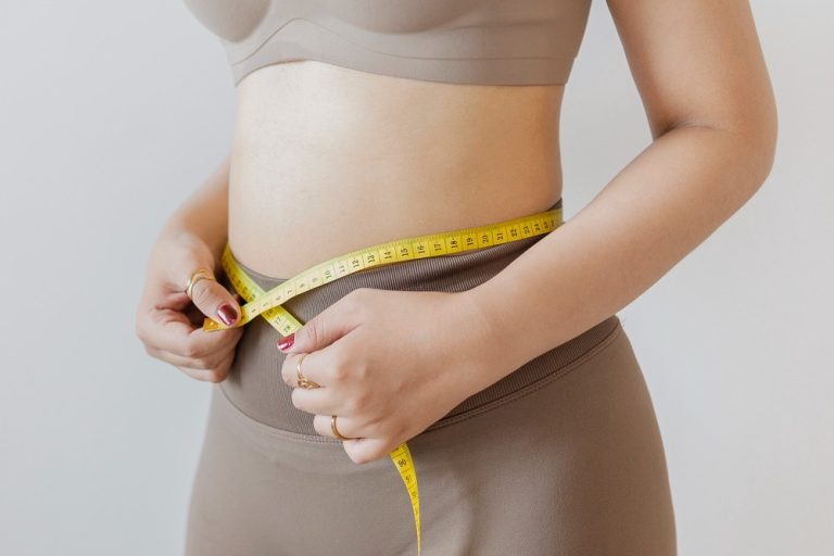 A woman in athletic wear measures her waistline with a tape measure after starting weight loss shots Houston.